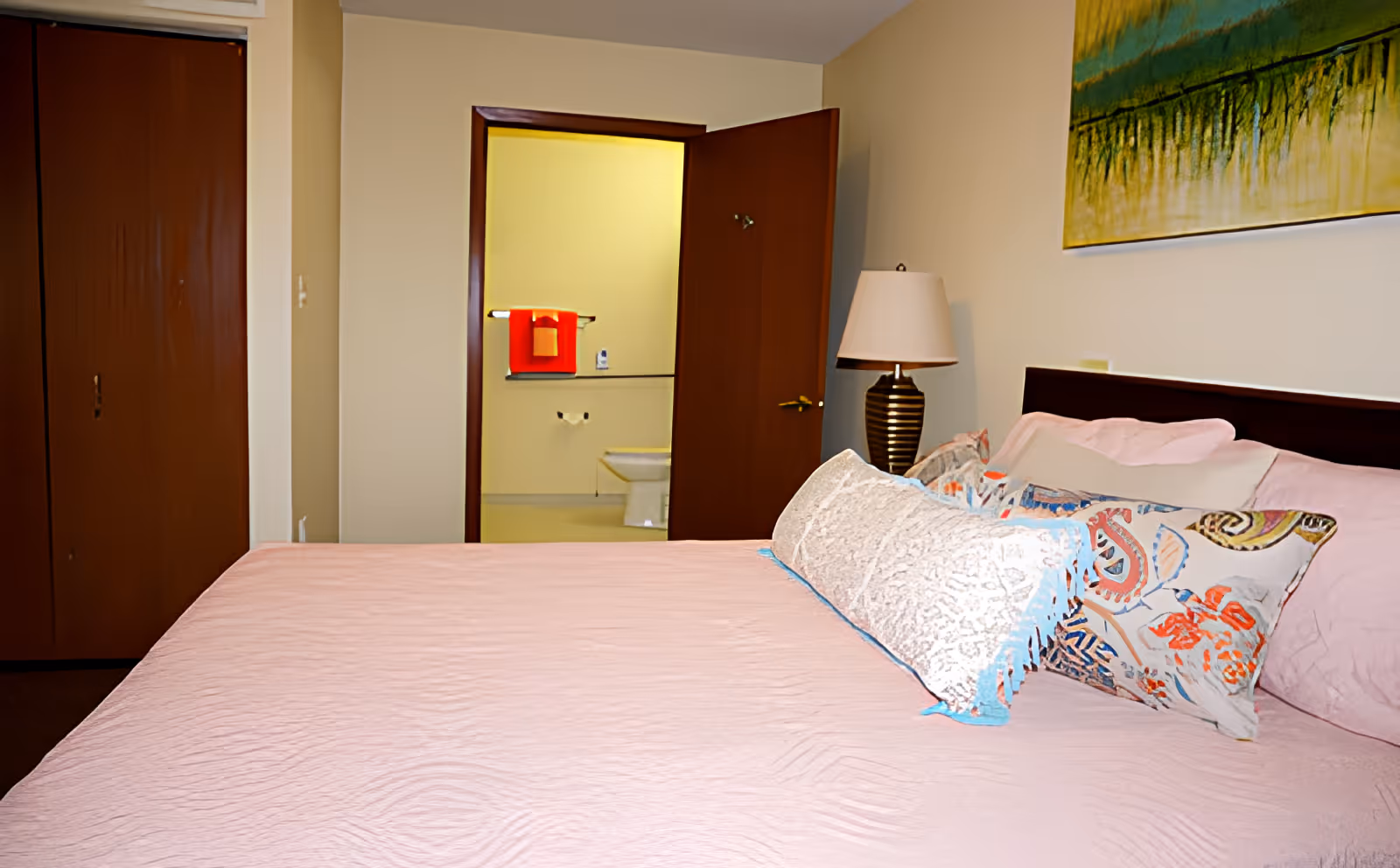 A bedroom with a large bed covered in a light pink bedspread and several decorative pillows. To the right of the bed is a bedside table with a lamp. On the wall above the bed is a colorful abstract painting. In the background, an open door reveals a bathroom with a toilet and a towel rack holding a red towel.