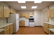 A clean kitchen with wooden cabinets, white refrigerator, white stove with oven, microwave, and a double sink. The floor is wooden, and the ceiling has recessed lighting panels.
