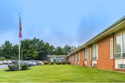 Exterior view of a single-story brick building with large windows, a green lawn in front, a flagpole with a flag, and a parking lot with several cars. Trees are visible in the background under a cloudy sky.