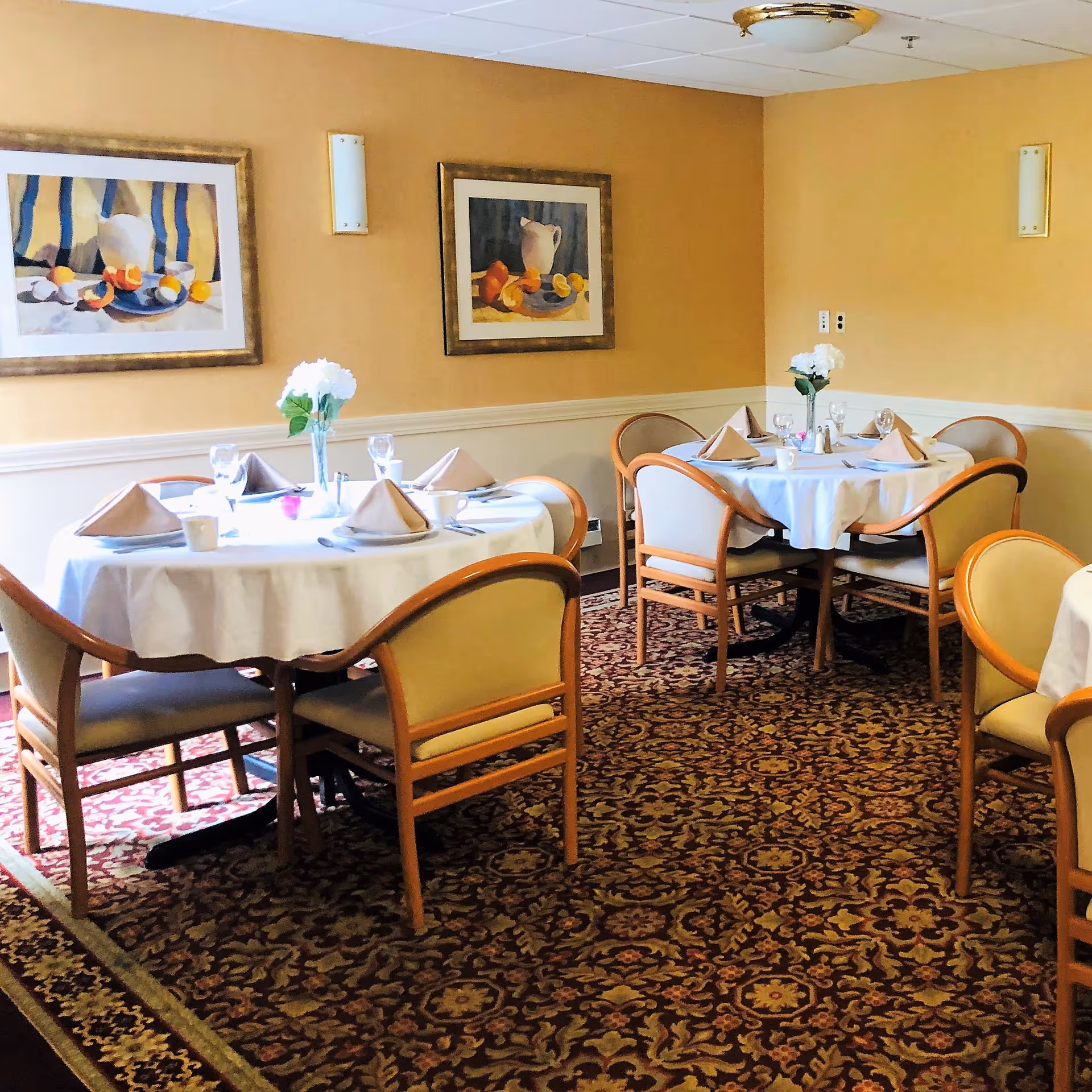 A dining room with round tables covered in white tablecloths, each set with folded beige napkins, glassware, and white flowers in vases. The room has patterned carpet, beige walls with two framed paintings of still life featuring fruit and pitchers, and wooden chairs with beige cushions.