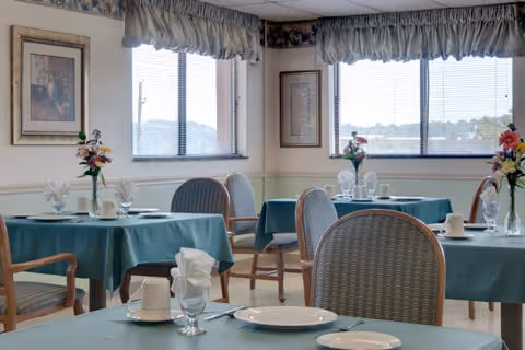 A dining room with multiple round tables covered with teal tablecloths, each set with white plates, cups, glasses, and napkins. The room has large windows with valance curtains, framed artwork on the walls, and wooden chairs with cushioned seats around the tables.