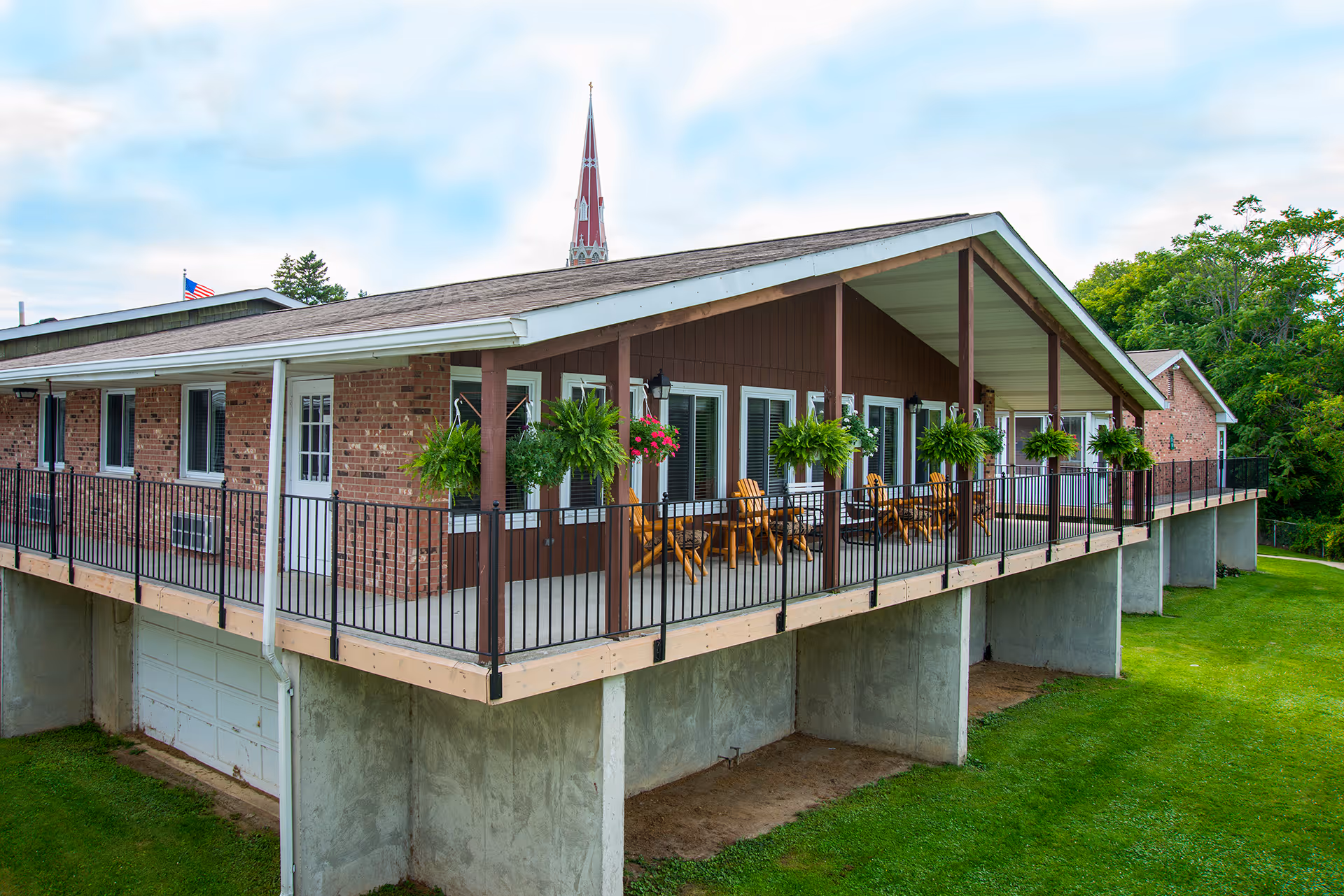 Exterior view of a raised brick and wood building with a covered porch featuring several wooden chairs and hanging plants. The building is surrounded by green grass and trees, with a church steeple visible in the background under a partly cloudy sky.
