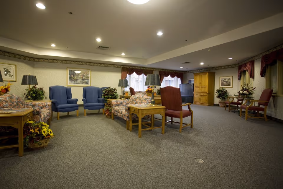 A spacious living room area in a senior living facility with multiple upholstered armchairs and sofas arranged around wooden side tables. The room is carpeted and decorated with framed artwork, potted plants, and table lamps. Large windows with red valances allow natural light into the room.