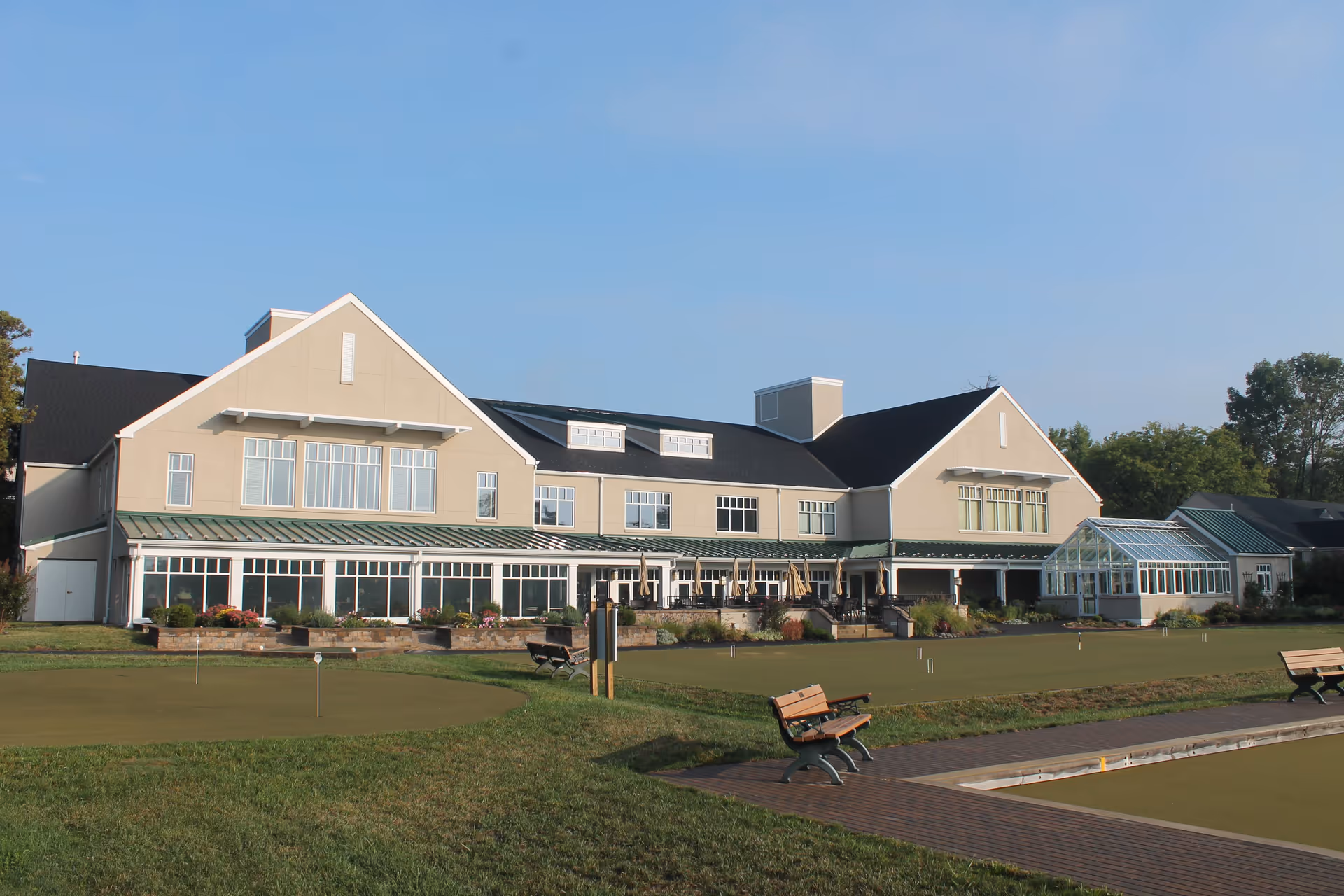 Exterior view of a large beige building with multiple windows and a black roof, surrounded by green lawns and a putting green area with benches and a small wooden walkway in the foreground under a clear blue sky.