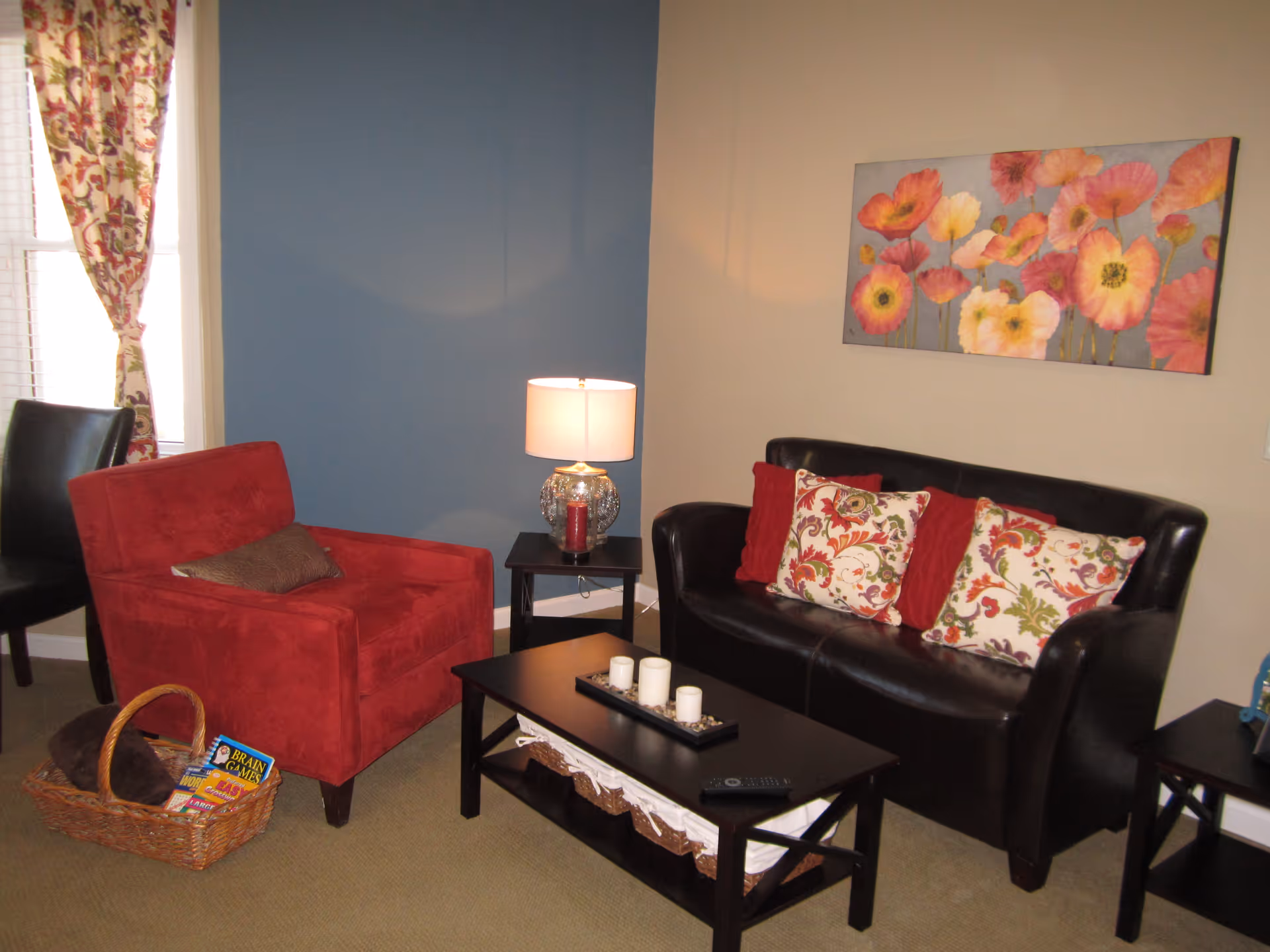 Cozy living room featuring a red armchair, black leather loveseat with floral pillows, a coffee table and side tables with a lamp against blue and beige walls.