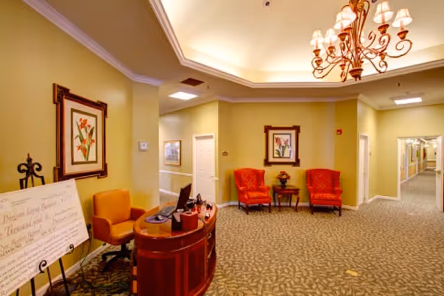 Interior view of a senior living facility hallway with beige walls and patterned carpet. There is a wooden reception desk with a computer and an orange chair on the left side. Two red upholstered chairs flank a small table with a flower arrangement against the far wall. Framed floral artwork hangs on the walls, and a decorative chandelier is mounted on the ceiling. Several doorways and a long corridor are visible in the background.