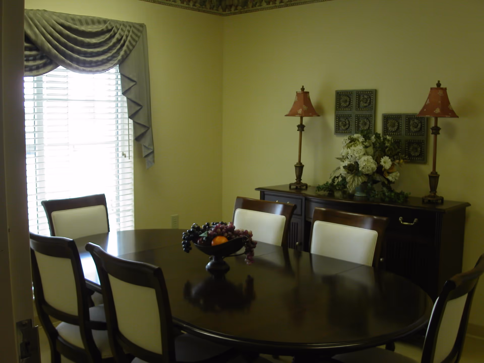 A dining room with a dark wooden oval table surrounded by six chairs with white cushions. On the table is a decorative bowl filled with artificial fruit. Against the wall is a dark wooden sideboard with two matching lamps and a floral arrangement. A window with white blinds and a draped valance is on the left side of the room.