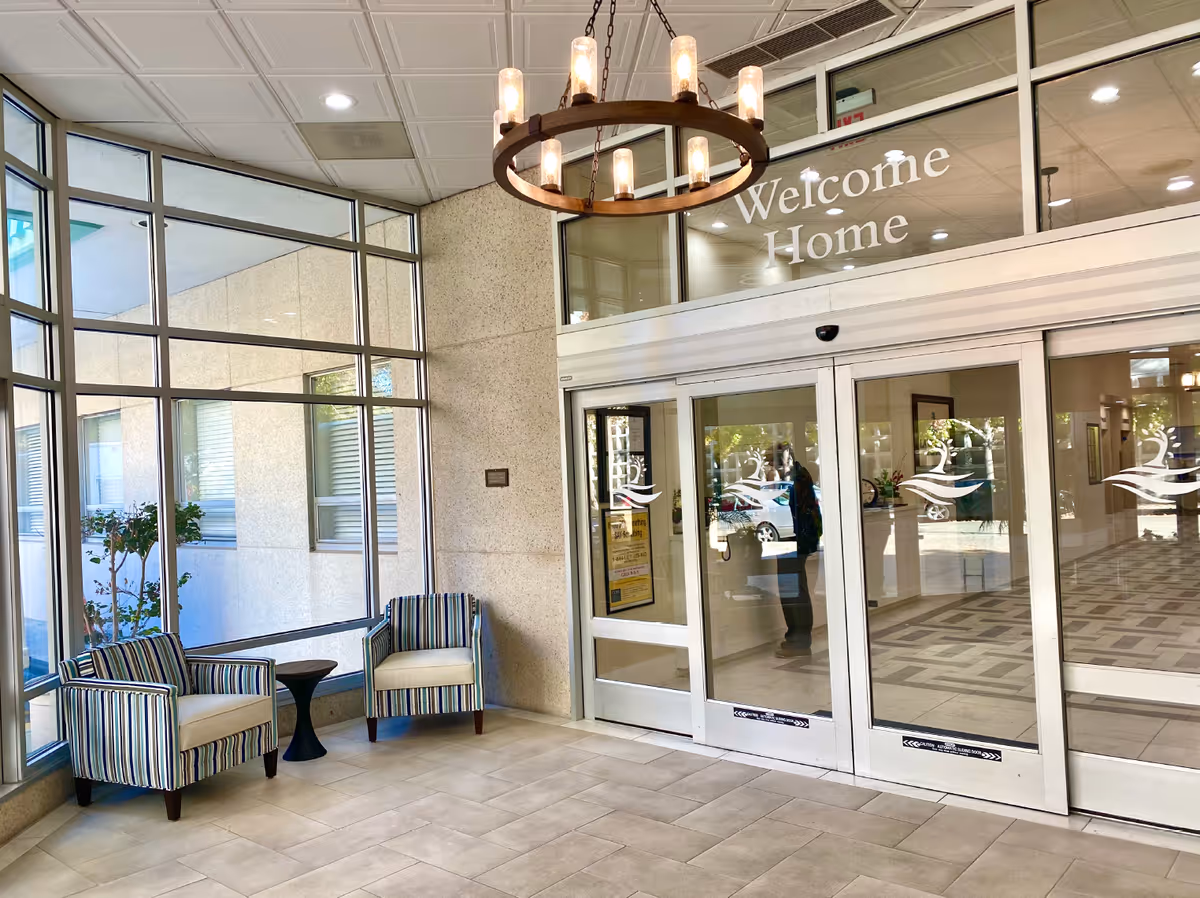 Entrance area of Lake Park Senior Living facility with large glass windows, two striped armchairs with a small black side table between them, a chandelier hanging from the ceiling, and automatic sliding glass doors with a 'Welcome Home' sign above.