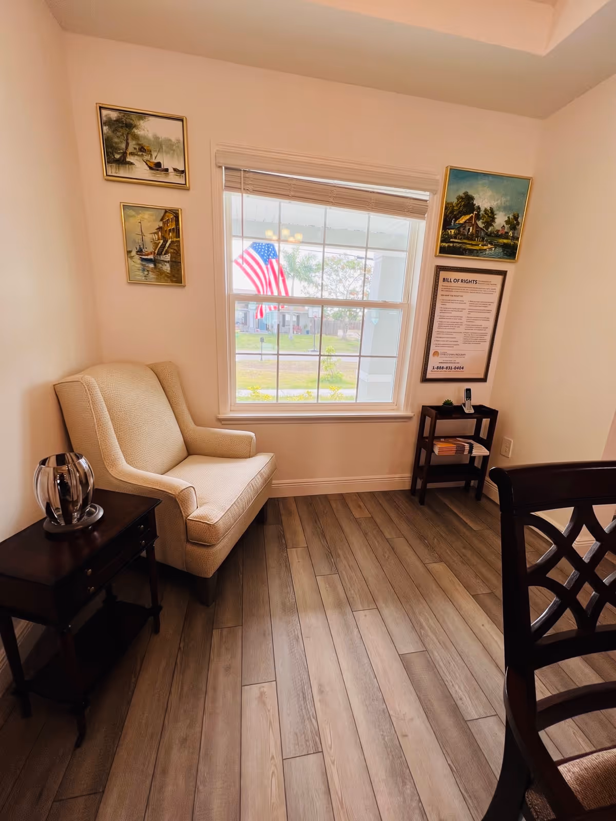 Cozy sitting area with an upholstered armchair beside a window showing an American flag, side tables, framed artwork, and wood-look flooring.