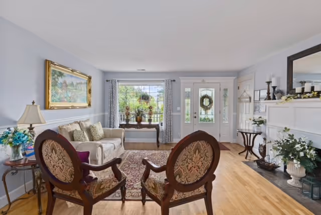 A bright living room with light-colored walls and hardwood floors. The room features a white sofa with decorative pillows, two ornate wooden armchairs with patterned upholstery, a wooden side table with a lamp and flowers, a large window with curtains, a front door with glass panels and a wreath, a fireplace with candles and floral decorations, and a large mirror above the fireplace.