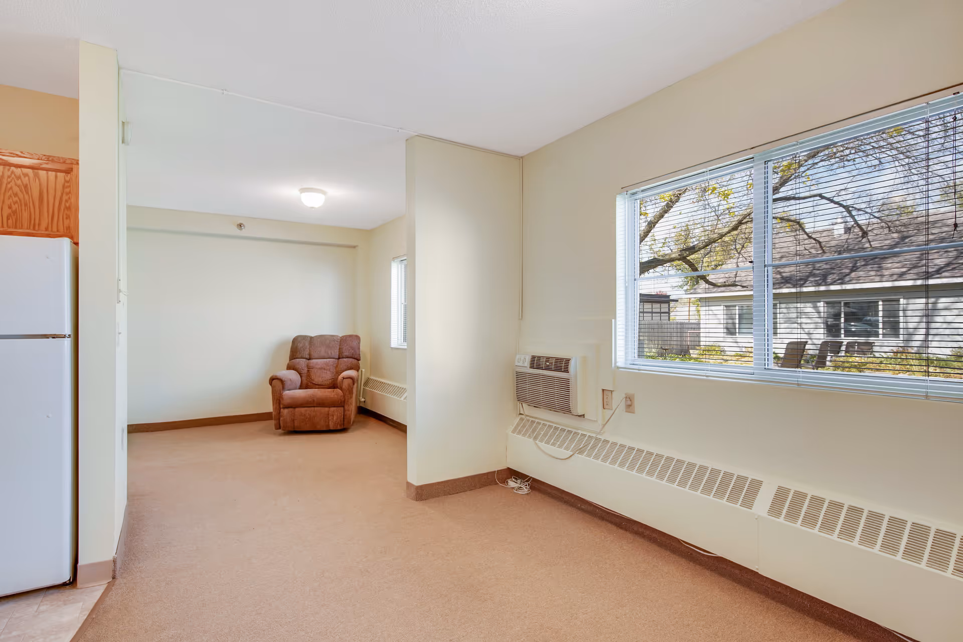 Interior view of a room in an assisted living facility with beige walls and carpet. There is a single brown recliner chair against the far wall, a white refrigerator partially visible on the left, a window with blinds on the right showing an outdoor view of trees and a neighboring building, and a wall-mounted air conditioning unit below the window.