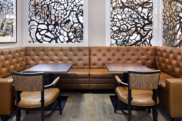 A cozy seating area with a brown tufted leather banquette along the wall, two wooden tables, and two cushioned chairs with patterned backs. Behind the seating are three windows with decorative black and white abstract patterned screens.