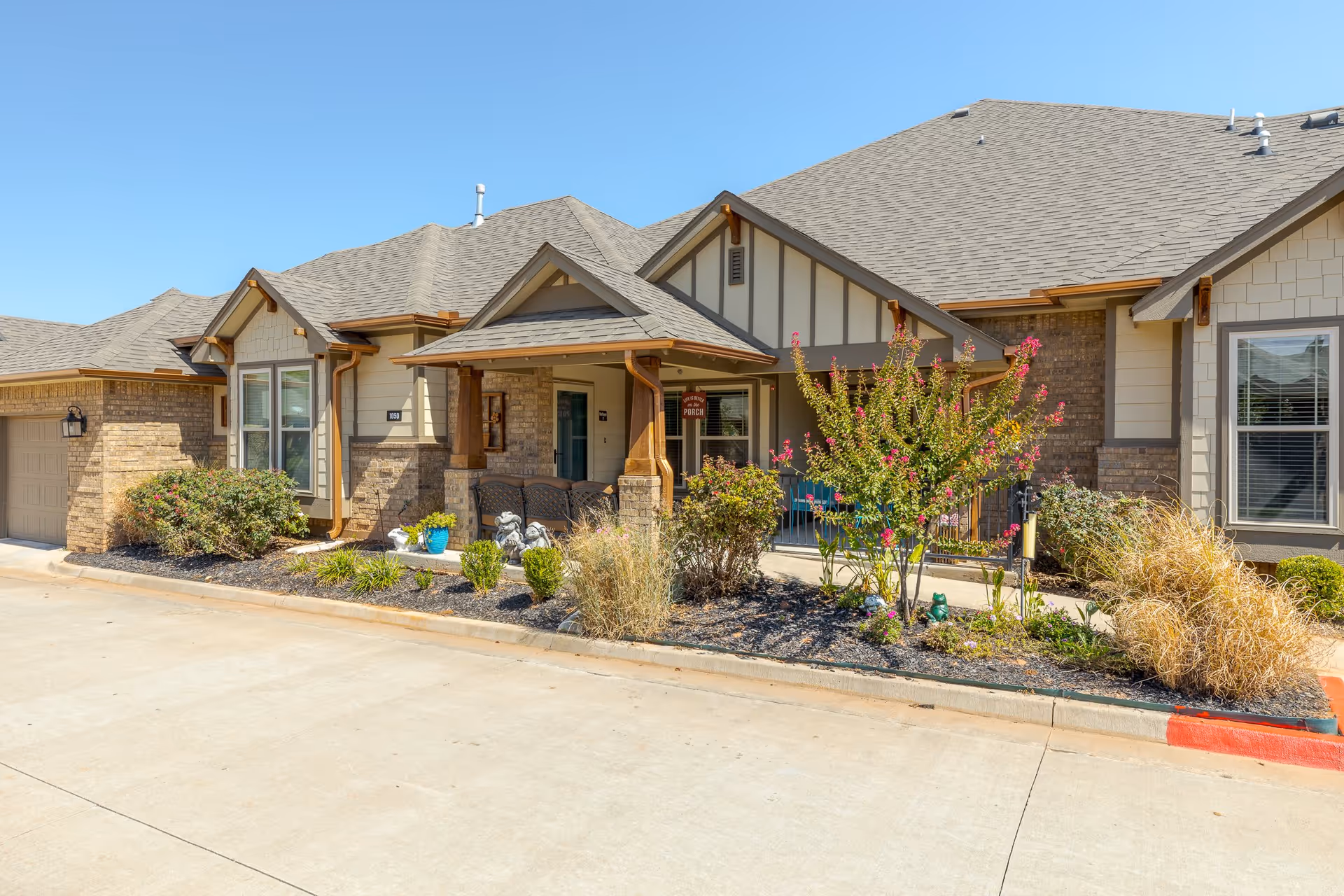 Exterior view of a single-story residential building with a covered porch, brick and siding walls, and a well-maintained garden with shrubs and flowering plants in front. The sky is clear and blue.