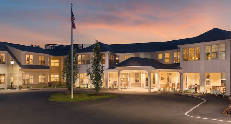Exterior front entrance of a two-story senior living facility at dusk with an American flag and illuminated windows.