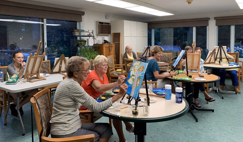 A group of elderly women seated around tables in a well-lit room, engaged in painting on canvases set on easels. The room has large windows with views of greenery outside and is furnished with wooden chairs and tables. Various painting supplies are visible on the tables.