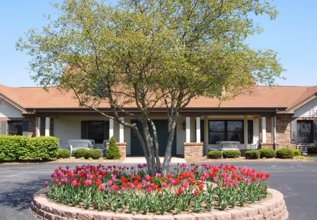 Single-story building facade with a central tree in a circular tulip flowerbed and porch seating under a clear blue sky.