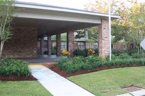Brick building entrance with a covered walkway, manicured landscaping, and a concrete sidewalk.