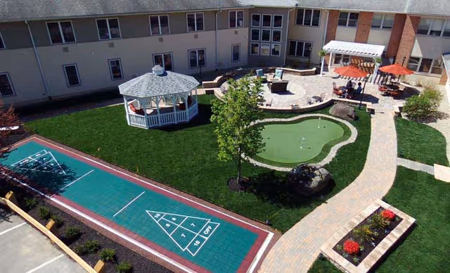 A landscaped outdoor courtyard at a senior living facility featuring a shuffleboard court, a putting green, a white gazebo, a paved walking path, flower beds, and seating areas with tables and umbrellas. The courtyard is surrounded by a two-story building.