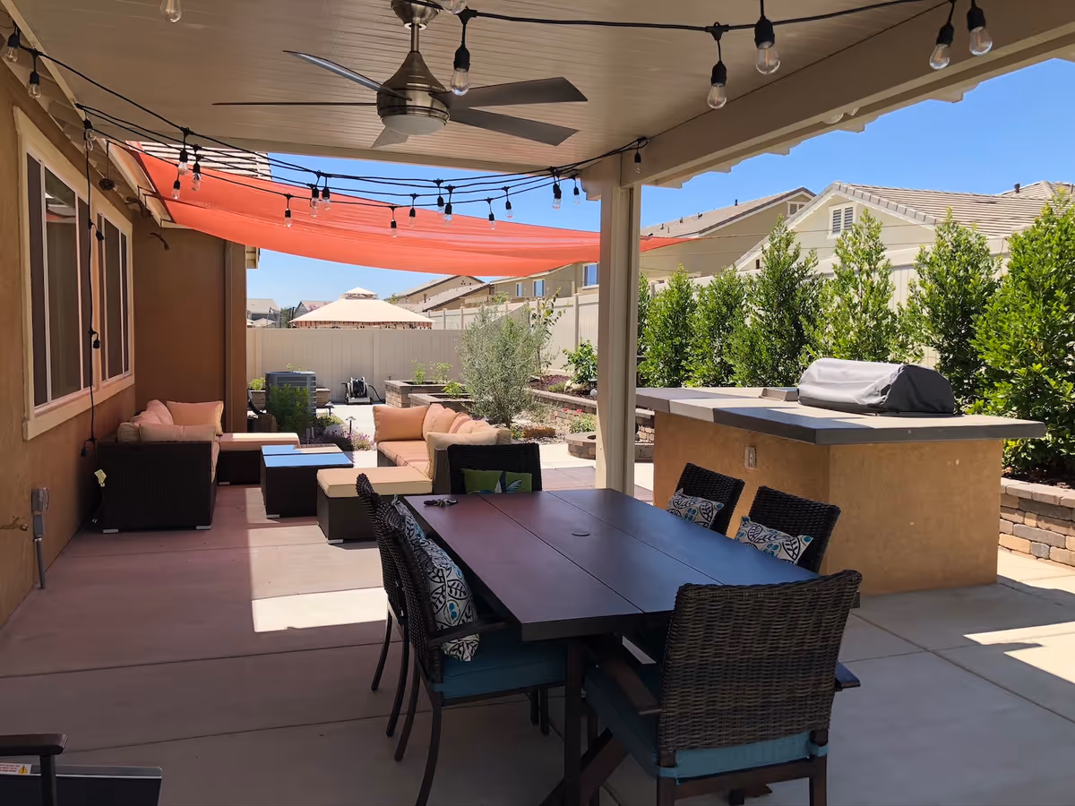 Covered outdoor patio area with a ceiling fan, string lights, and a red shade sail. There is a dining table with six wicker chairs featuring patterned cushions, a built-in barbecue grill, and a seating area with cushioned sofas and a coffee table. The patio overlooks a fenced backyard with green shrubs and neighboring houses under a clear blue sky.