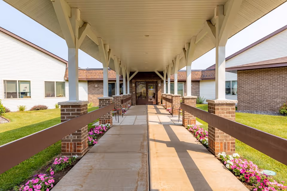 Covered walkway with brick pillars and wooden railings leading to the entrance of a building. The walkway is flanked by flower beds with pink flowers and green grass on both sides. The building has white siding and brick walls with windows visible on either side.