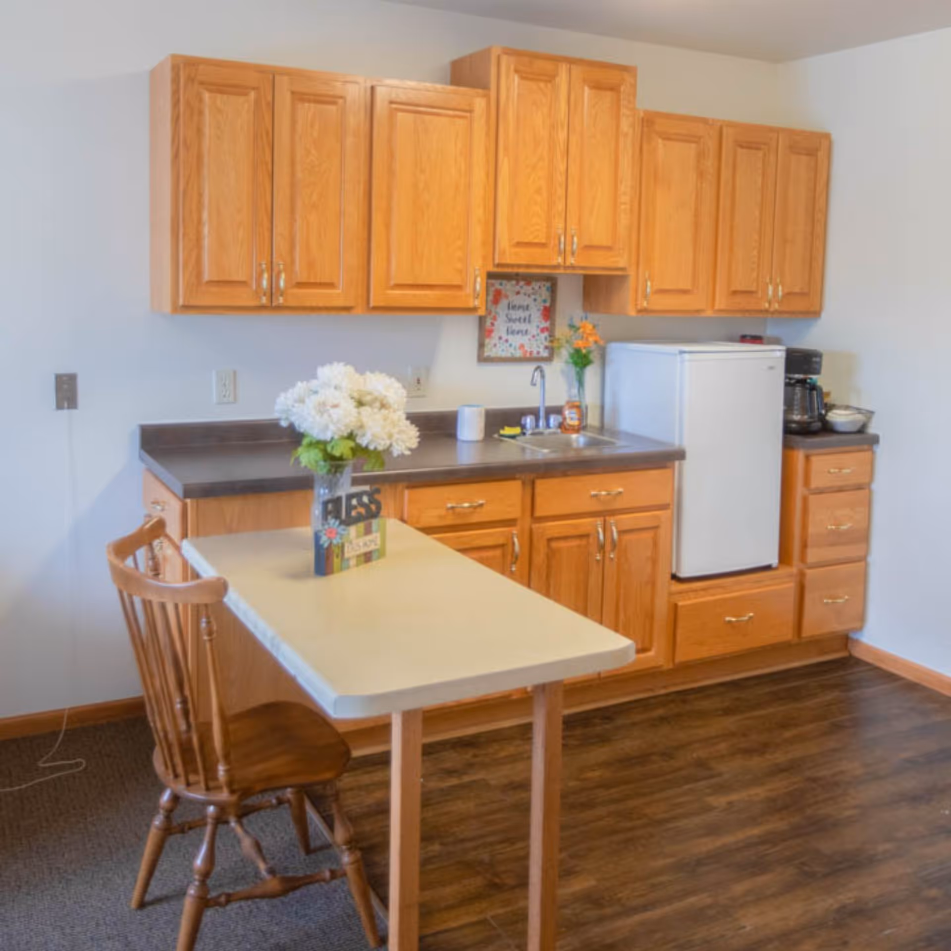 A small kitchen area with wooden cabinets, a countertop with a sink, a white mini refrigerator, a coffee maker, and a small table with a wooden chair. On the table is a vase with white flowers and a decorative sign that says 'Bless this home'.