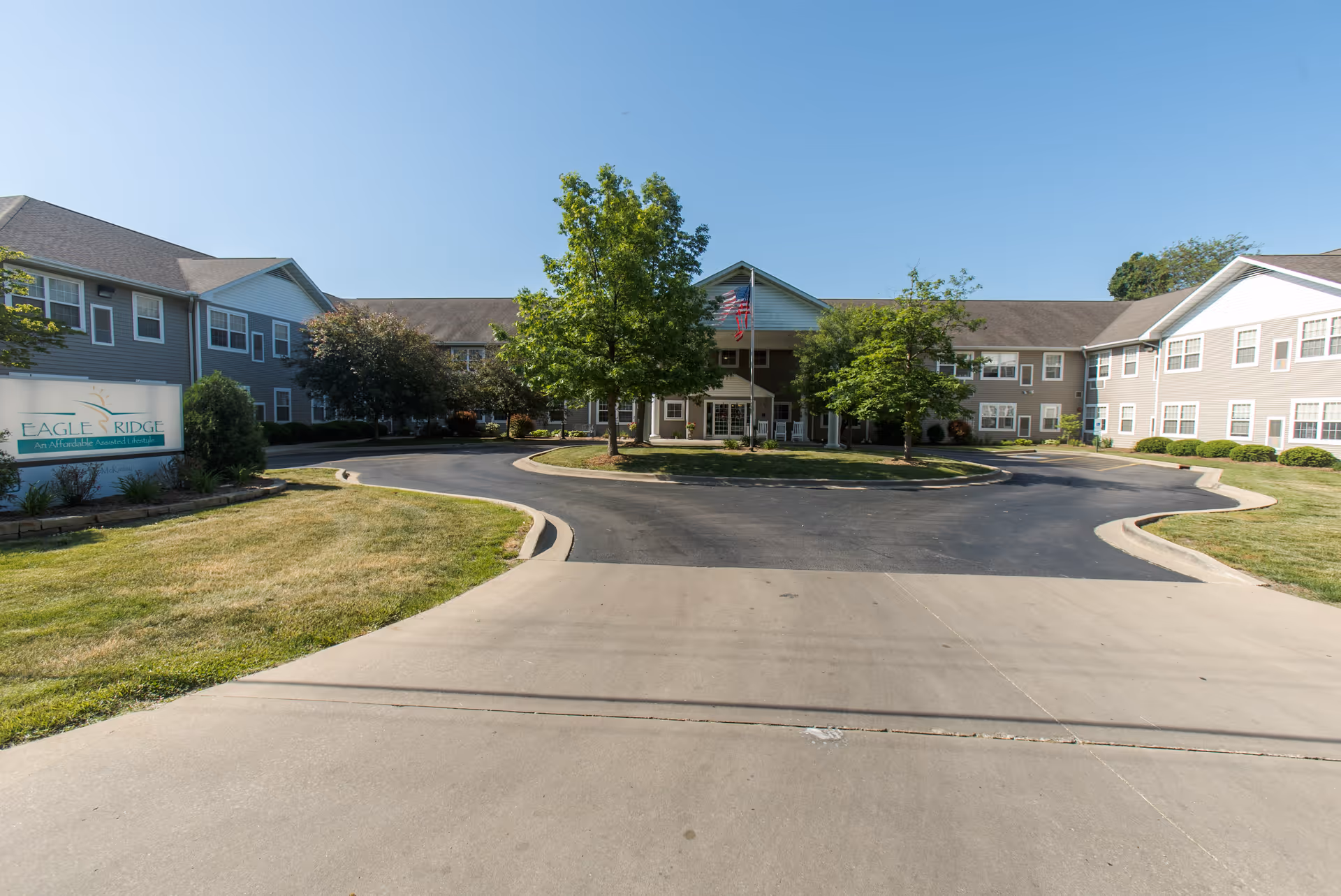 Front exterior view of Eagle Ridge of Decatur, a two-story residential building with gray siding and white trim. The building is U-shaped with a driveway and circular entrance in front, surrounded by green lawns and trees. An American flag is displayed near the entrance, and a sign on the left reads 'Eagle Ridge An Affordable Assisted Living'.