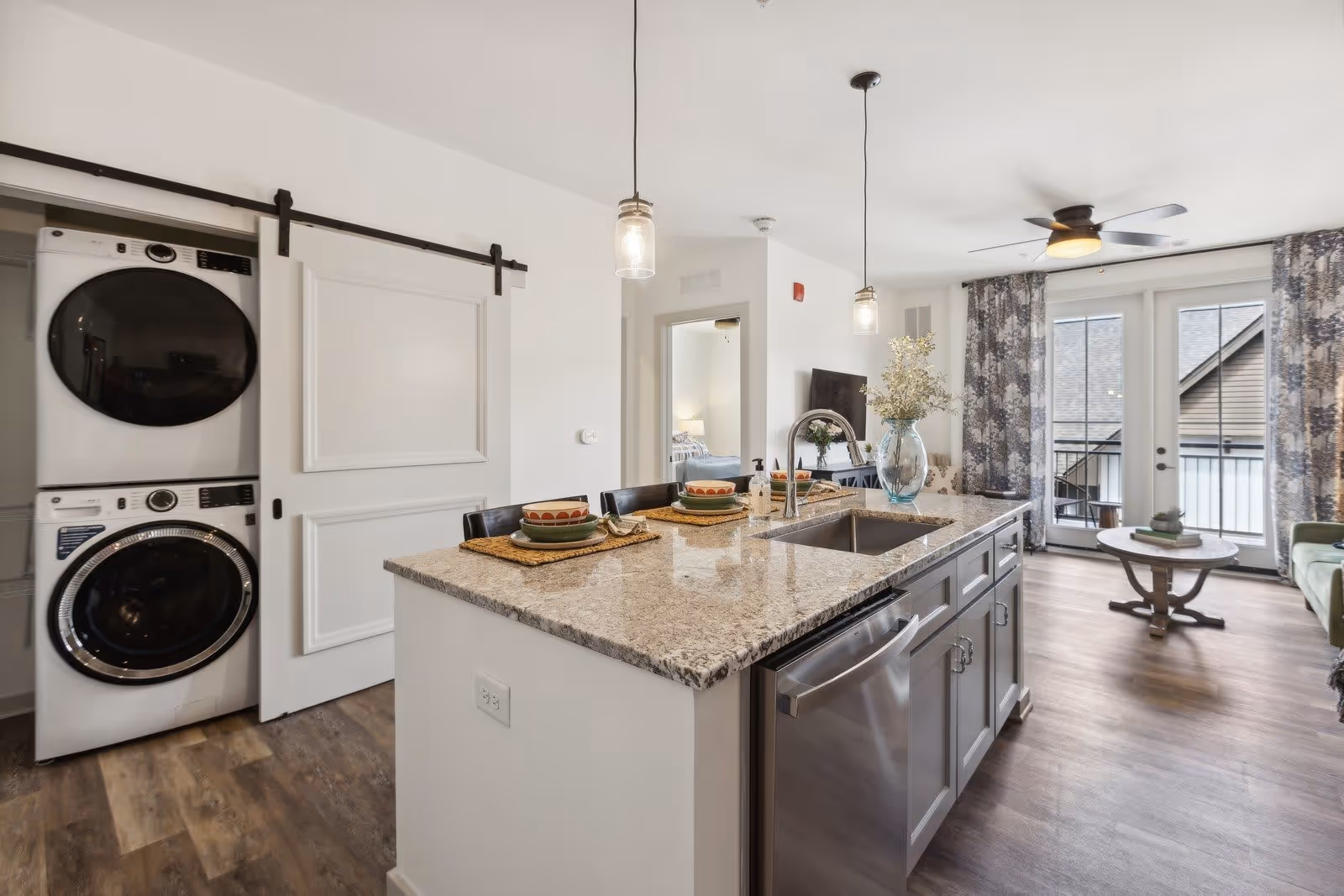 Modern open-concept living space featuring a kitchen island with granite countertop, two pendant lights, a built-in sink, and place settings for two. To the left, there is a stacked washer and dryer behind a sliding barn door. In the background, a living area with a ceiling fan, floral curtains, a round coffee table, and glass doors leading to a balcony is visible. A bedroom can be seen through an open doorway.