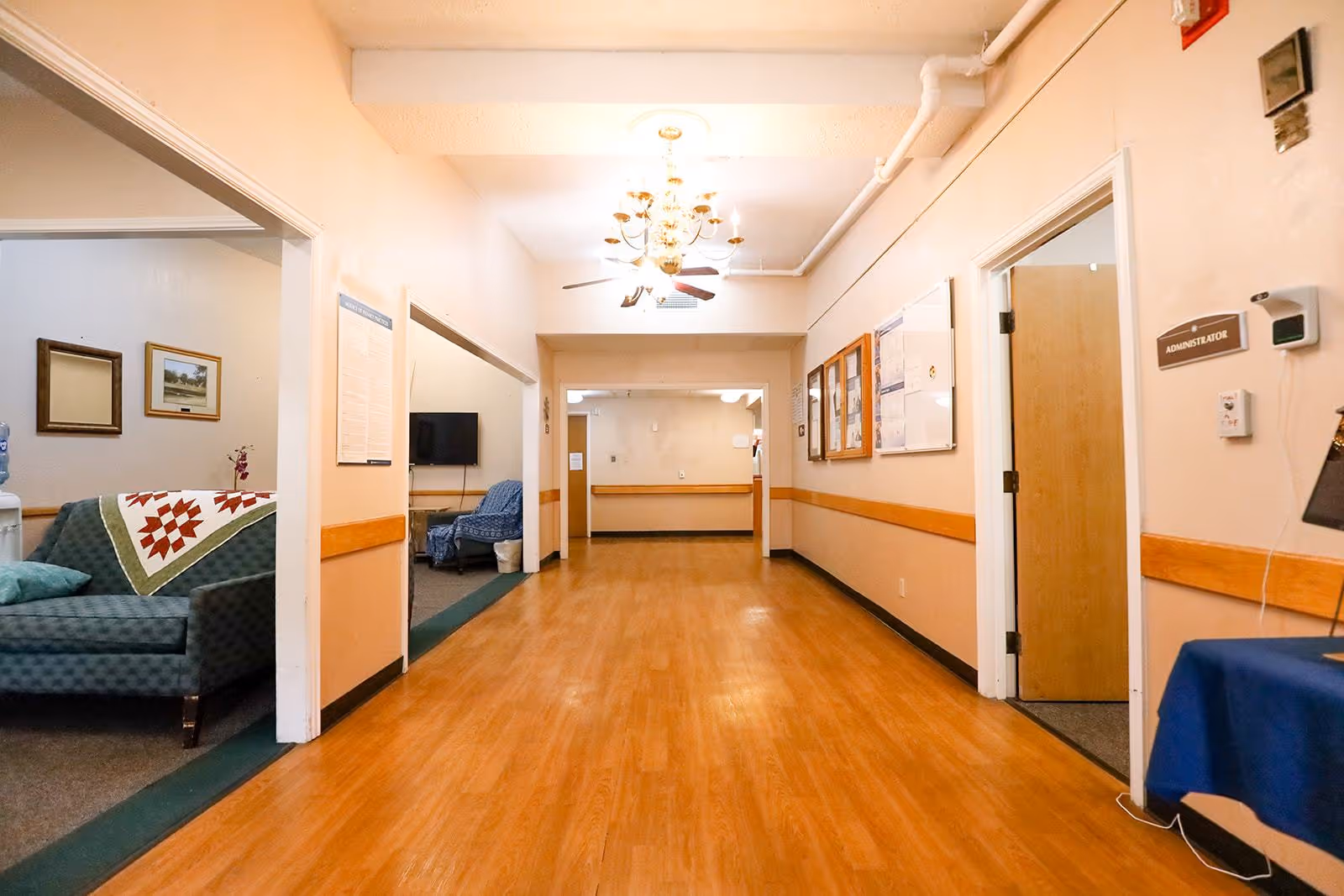 A wide hallway with wooden flooring and beige walls in a healthcare facility. On the left, there is a seating area with a green couch covered by a quilt and a blue armchair, along with framed pictures on the wall. On the right, there is a door labeled 'Administrator' and bulletin boards with notices. The ceiling has a chandelier and ceiling fans, and the hallway extends into another room in the background.