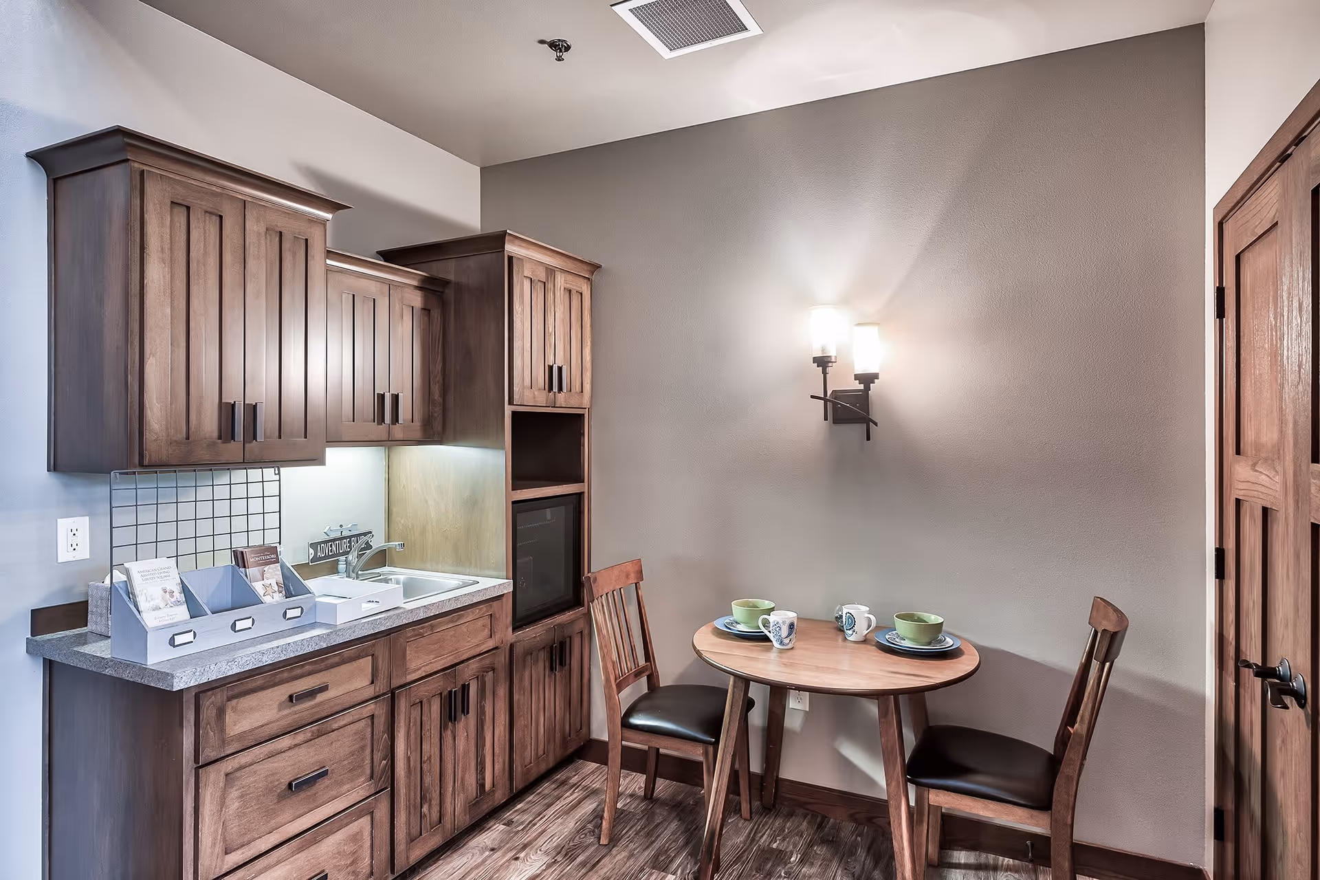 A small kitchenette area with wooden cabinets, a sink, and a countertop. Next to the kitchenette is a round wooden table set with two chairs, each with a place setting including a plate, bowl, and mug. A wall-mounted light fixture illuminates the space, and there is a wooden door to the right.