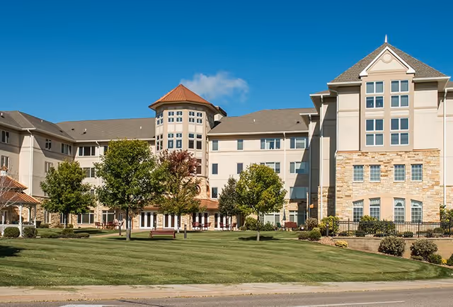 Exterior view of a large senior living facility building with multiple windows, a central tower with a conical roof, and a well-maintained lawn with trees and benches in front under a clear blue sky.