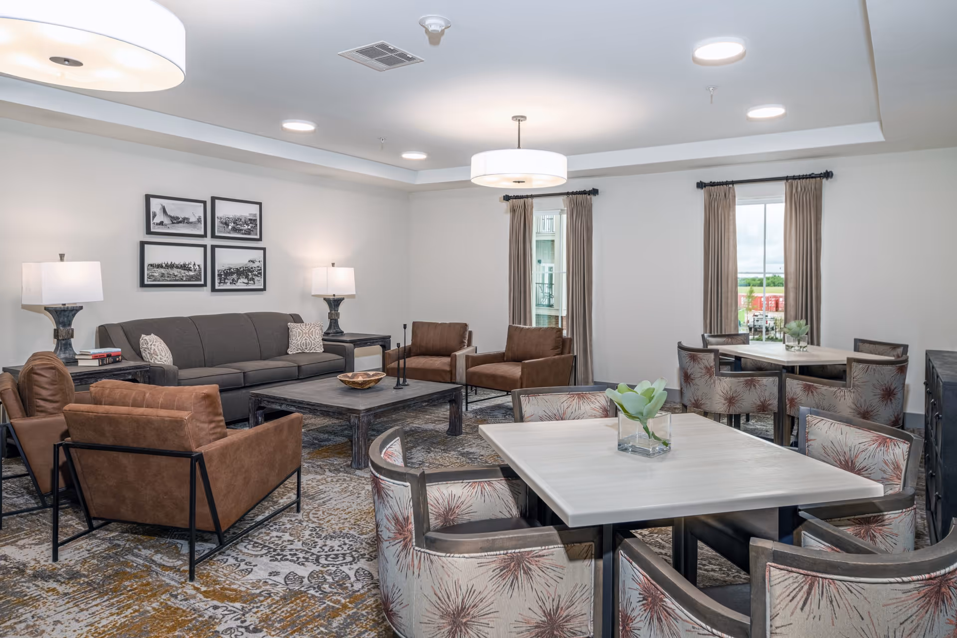 A well-lit senior living common area featuring a gray sofa, four brown armchairs, two square coffee tables, and two dining tables with patterned chairs. The room has two windows with beige curtains, framed black and white photos on the wall, and modern ceiling lights.