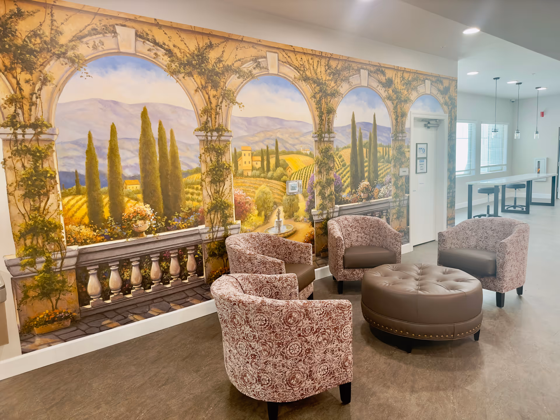 Seating area with four patterned armchairs and a round leather ottoman in front of a large mural of an Italian-style countryside.