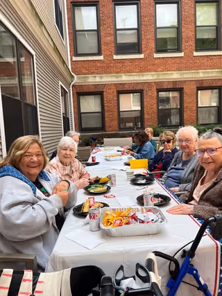 A group of elderly people sitting around a long outdoor table covered with a white tablecloth, enjoying a meal together. The table has trays of snacks, Diet Coke cans, and plates with food. The setting is in a courtyard surrounded by brick and siding buildings with multiple windows.