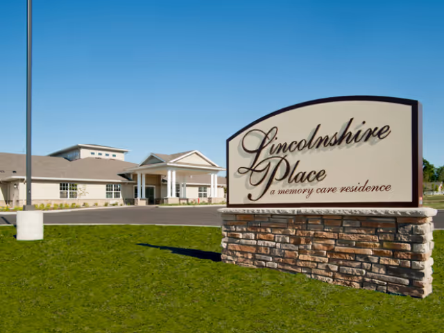 Exterior view of Lincolnshire Place, a memory care residence, showing the building and a large sign with the facility's name on a stone base, under a clear blue sky.