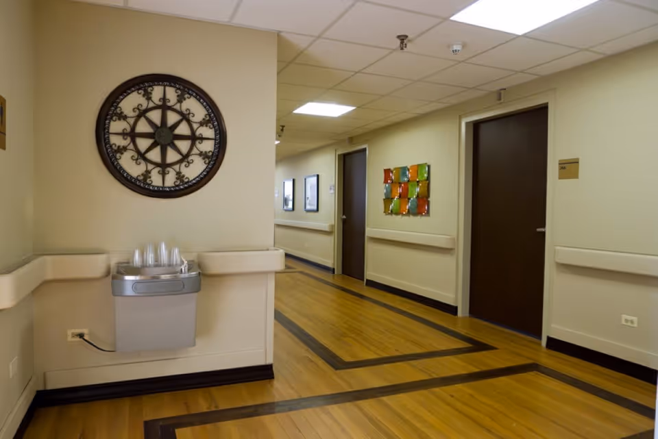 Well-lit interior hallway of a senior living facility with wooden floors, a wall-mounted water fountain, decorative wall art, and several closed doors.