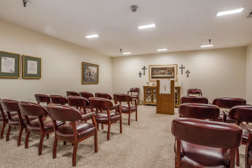 A small chapel or prayer room with rows of wooden chairs with red cushions facing a wooden podium adorned with a white cloth featuring a gold cross. The back wall has religious decorations including three crosses and a framed picture of The Last Supper. The room has beige walls, carpeted floor, and ceiling lights.