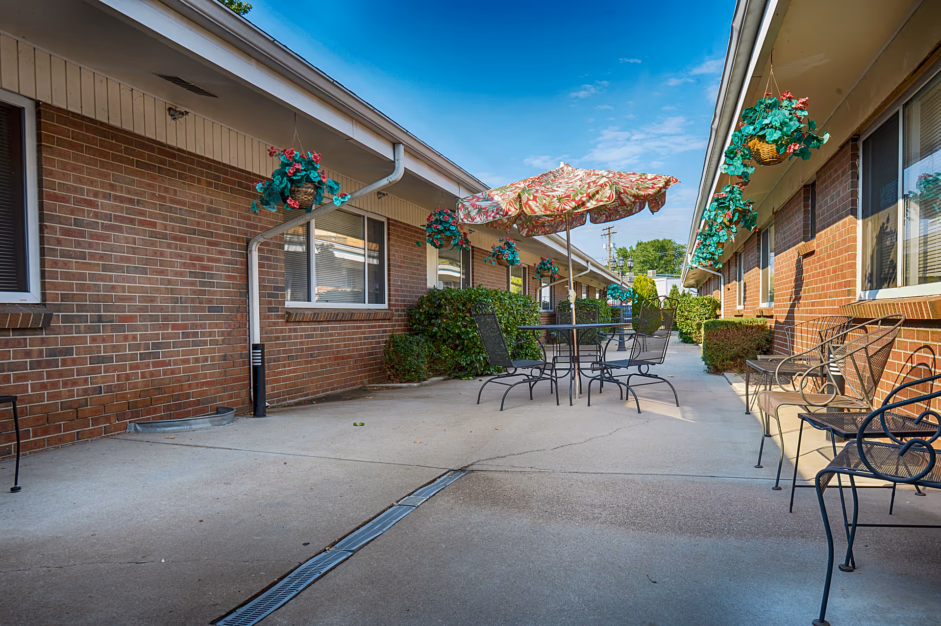 Outdoor patio area between two brick buildings with hanging flower baskets, metal chairs, and a table with a floral umbrella under a clear blue sky.