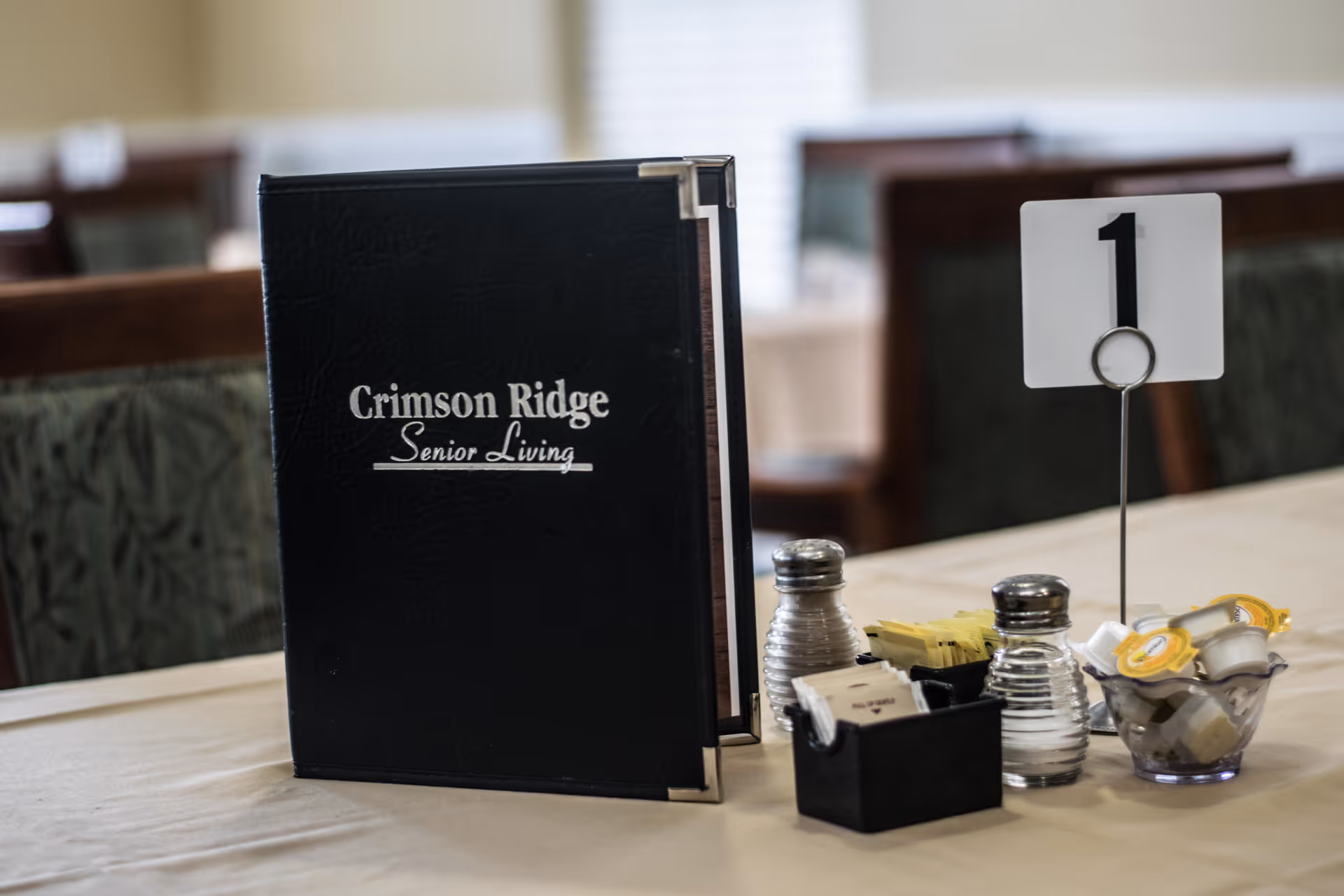 A dining table set with a black menu folder labeled 'Crimson Ridge Senior Living', a table number holder displaying the number 1, salt and pepper shakers, packets of sugar and sweeteners, and small containers of creamers in a senior living community dining room.