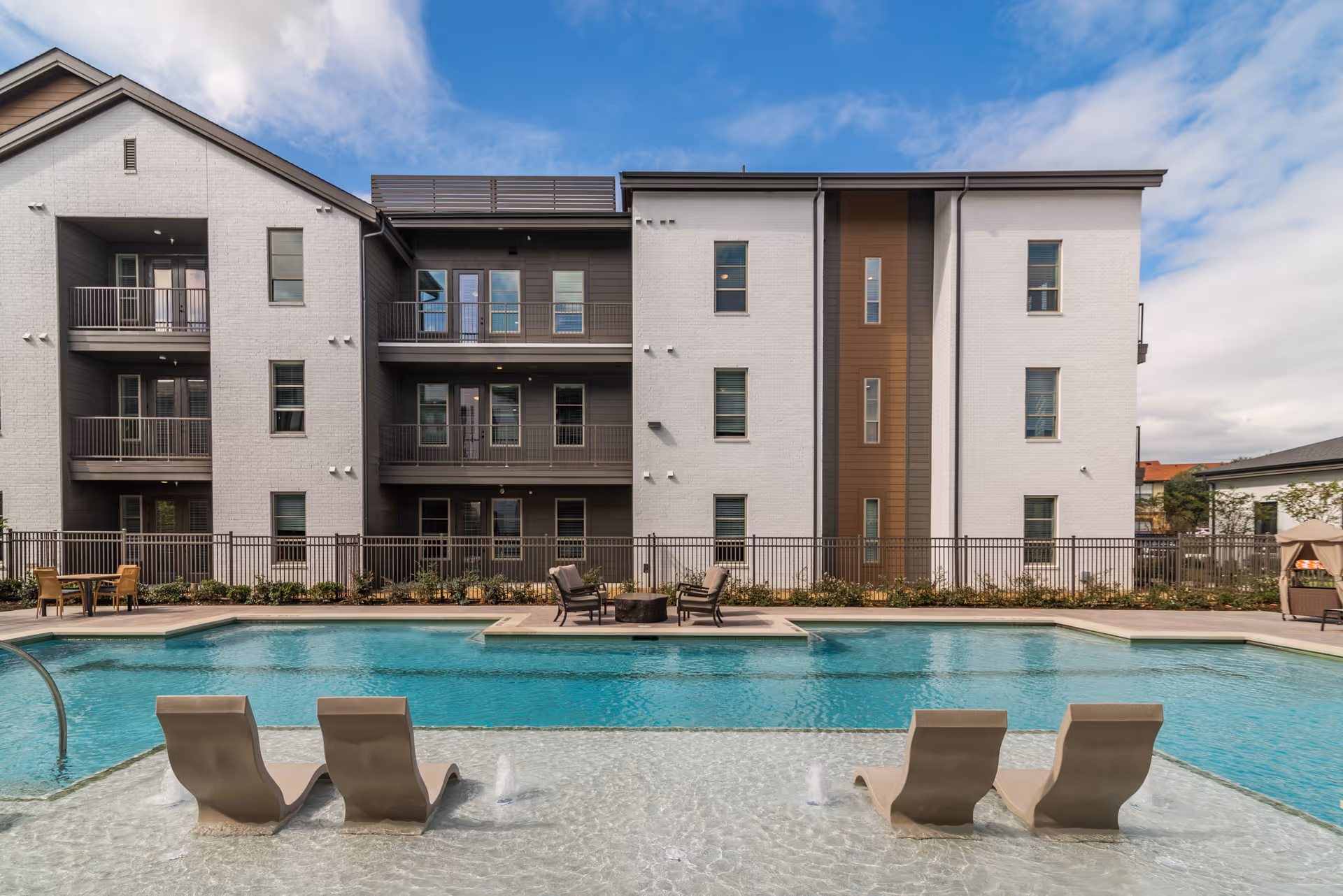 Outdoor swimming pool area with four lounge chairs partially submerged in shallow water, a fire pit with chairs on a raised platform in the middle of the pool, and a three-story residential building with balconies in the background under a partly cloudy sky.