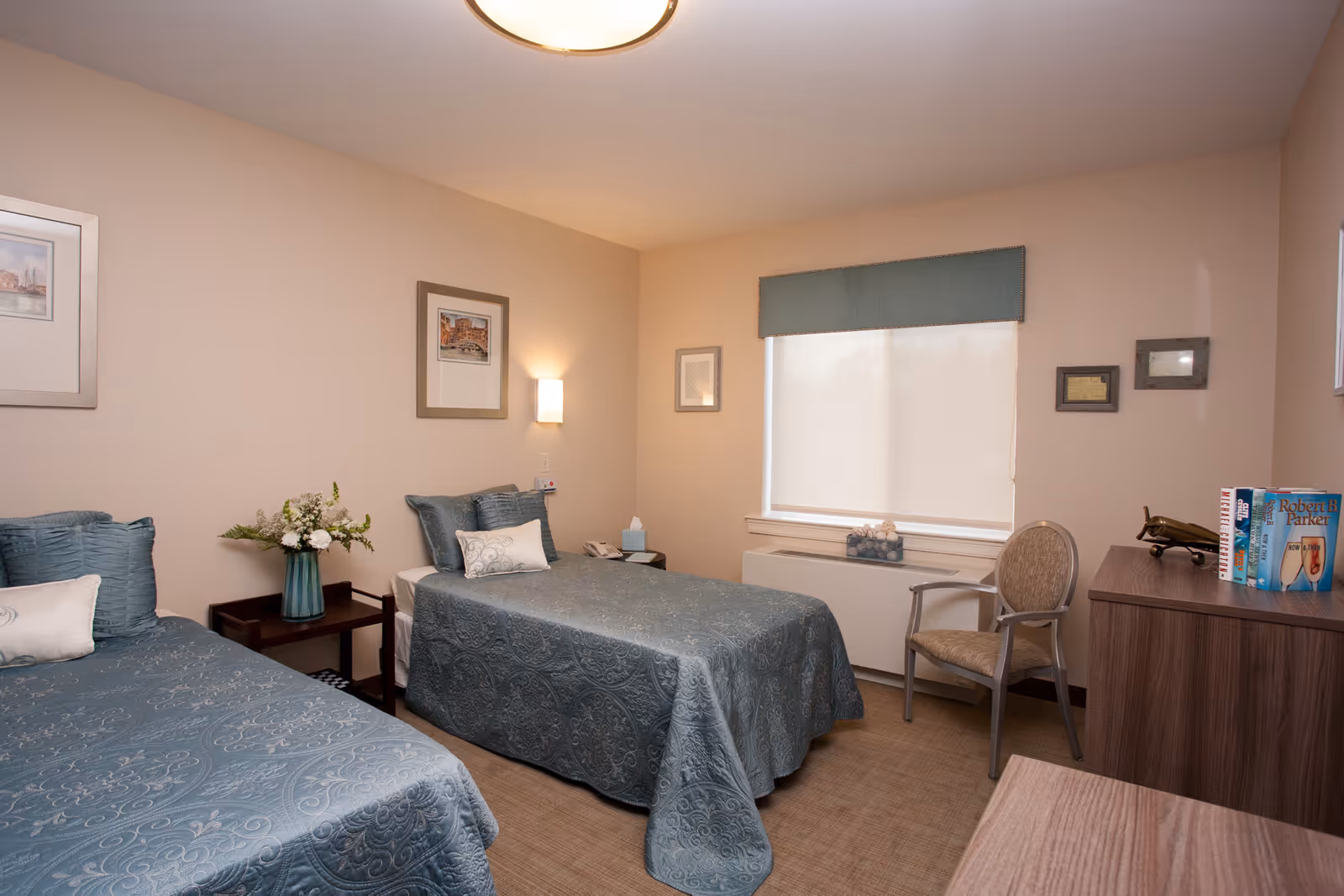 A cozy bedroom with two twin beds covered in matching blue patterned bedspreads. Between the beds is a small wooden nightstand with a vase of flowers. The room has beige walls adorned with framed pictures and a window with a blue valance and closed blinds. There is a wooden dresser with books and a decorative item on top, and a cushioned chair next to the window.