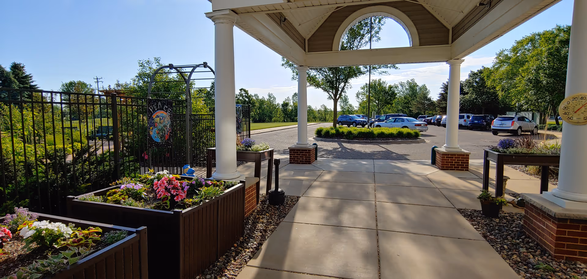 View from a covered entrance area with white columns and brick bases looking out onto a parking lot with several cars. Raised garden beds with colorful flowers are on the left side, and a black metal fence with a decorative sign is visible. Trees and greenery surround the parking area under a clear blue sky.