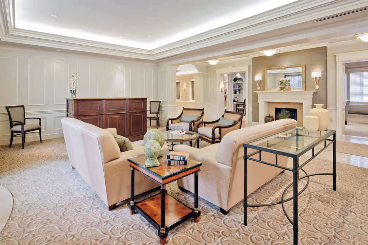 A spacious and elegant living room area in a senior living facility featuring two beige sofas arranged around a glass coffee table, several armchairs, a wooden reception desk, a fireplace with a mirror above it, and decorative vases on side tables. The room has soft lighting, carpeted floors, and white paneled walls.