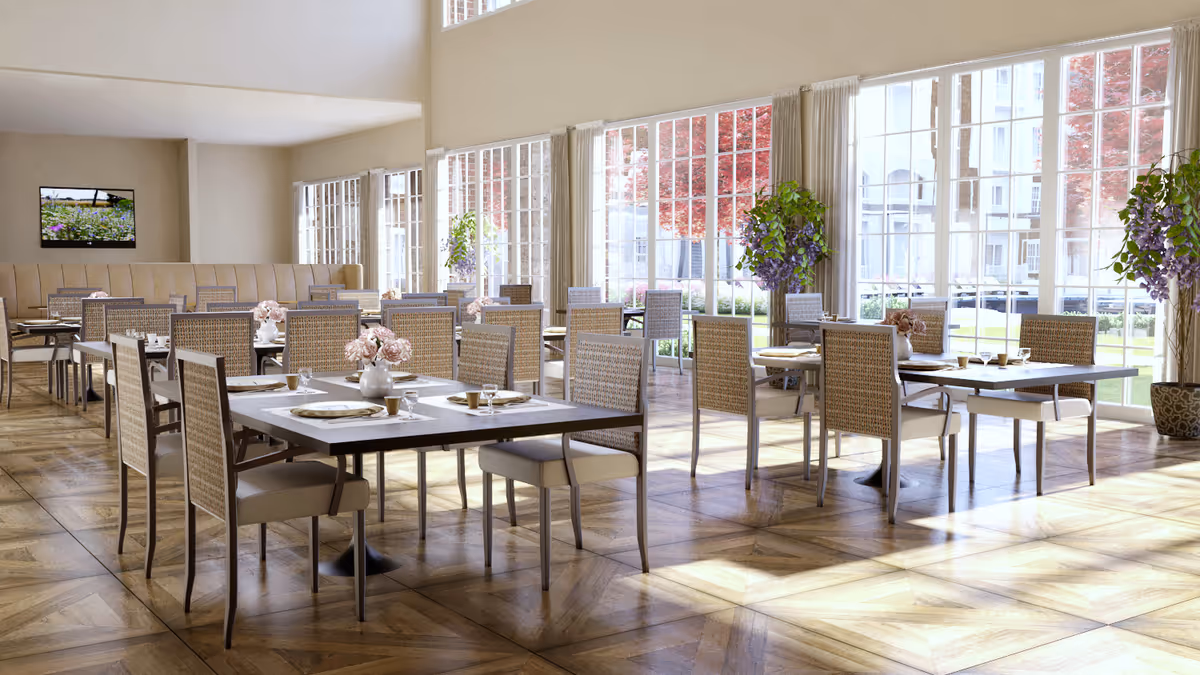 A bright and spacious dining room with multiple tables and chairs arranged neatly. Each table is set with plates, cups, and a vase of pink flowers. Large windows along one wall allow natural light to flood the room, revealing a view of greenery and red-leaved trees outside. The floor has a patterned wooden design, and there are potted plants with purple flowers near the windows. A flat-screen TV is mounted on the far wall above a beige cushioned bench.