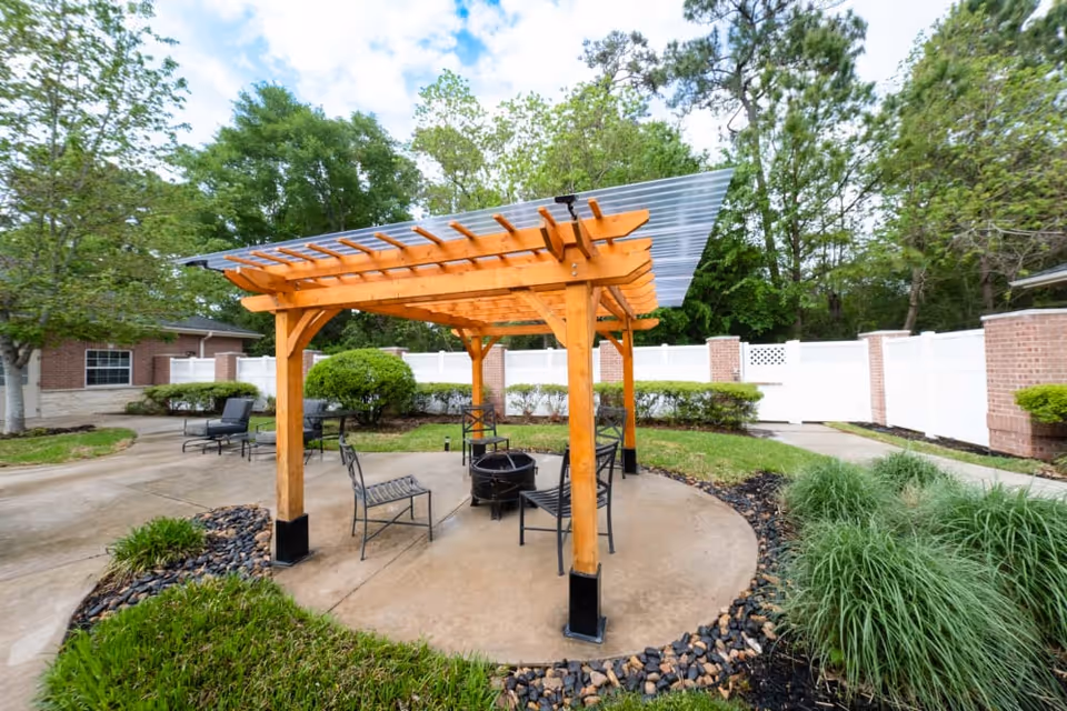 Wooden pergola with chairs around a fire pit in a landscaped courtyard with a white fence and trees.