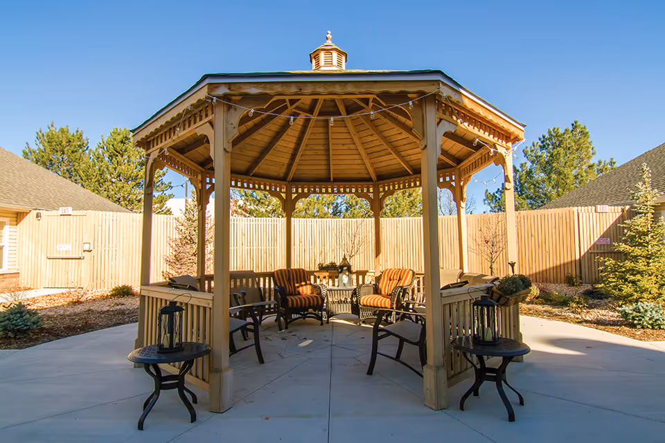 Outdoor wooden gazebo with a green roof and string lights, surrounded by a concrete patio. Inside the gazebo are cushioned chairs with striped upholstery and a small table with decorative lanterns. The area is enclosed by a wooden fence with trees and shrubs around.