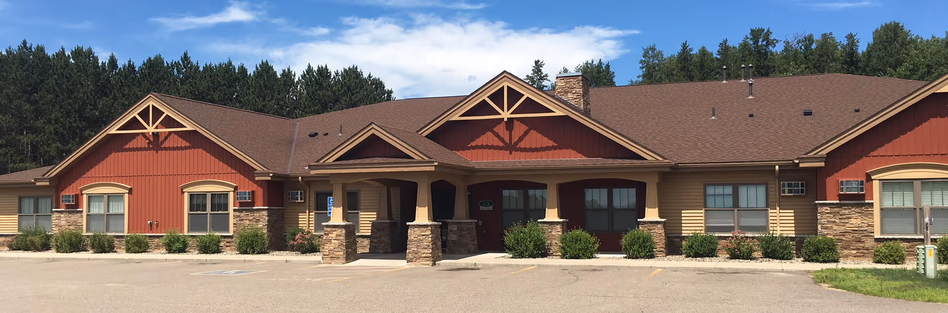 Front exterior view of a single-story assisted living facility building with a brown roof, red and beige siding, stone accents, and a covered entrance. There are several windows and some landscaping with bushes in front of the building. The sky is clear with a few clouds and there are trees in the background.
