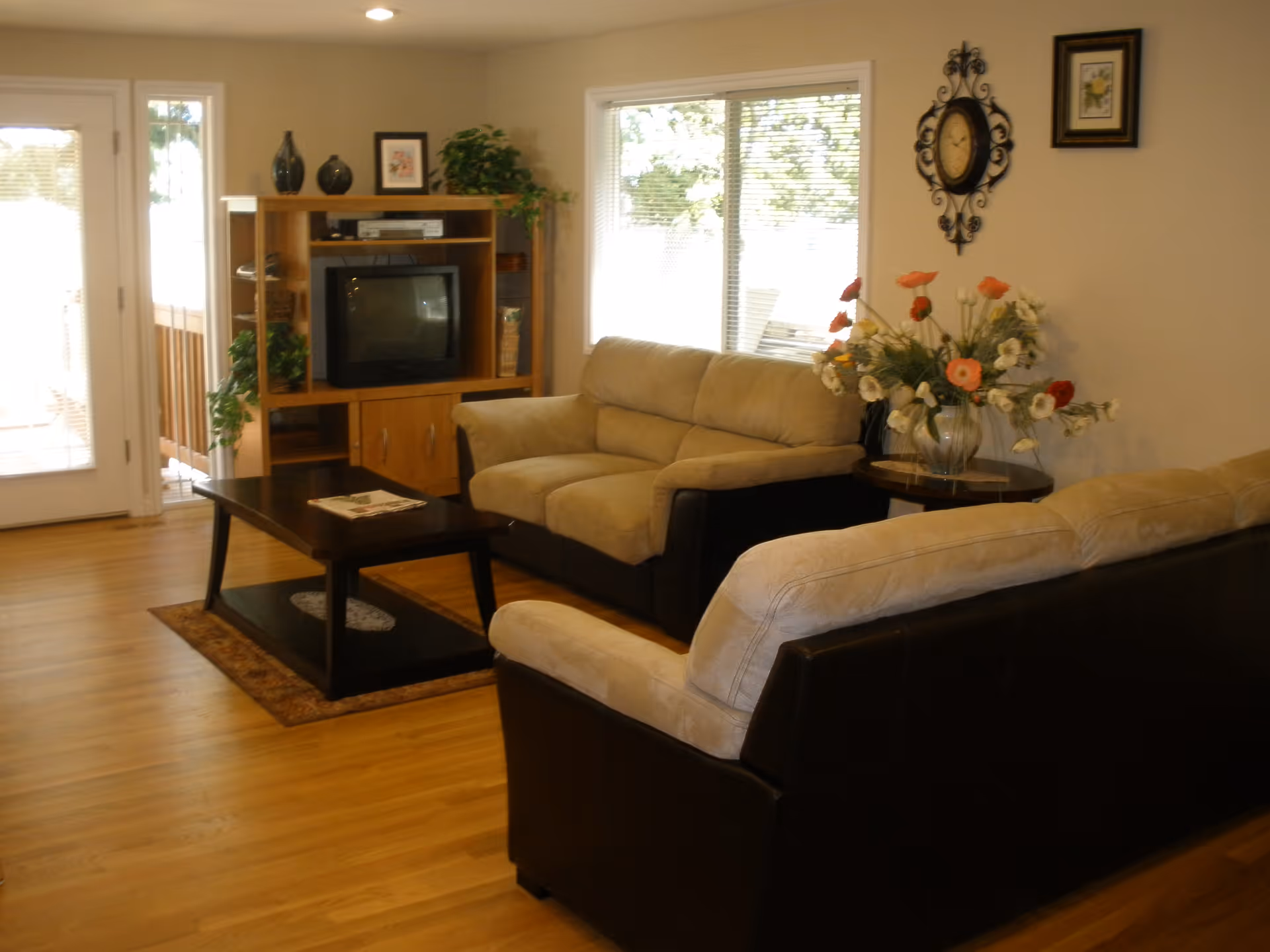 A cozy living room with two beige and brown sofas facing a wooden coffee table and an entertainment center holding a TV and decorative items. There is a large window letting in natural light, a wall clock, framed pictures, and a vase with flowers on a side table.