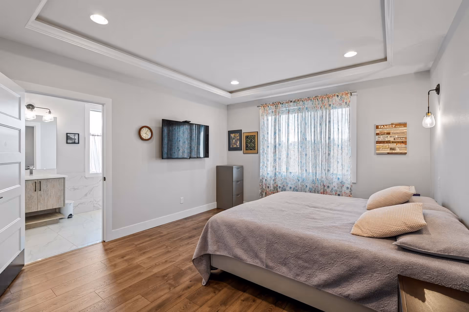 A bright bedroom with a large bed covered in a light gray quilt and two beige pillows. The room has wooden flooring, a wall-mounted flat-screen TV, a small gray dresser, and a window with floral sheer curtains. A door on the left leads to a bathroom with white marble tiles and a vanity with a mirror. The walls are painted light gray, and there is a wall clock and framed pictures on the walls.