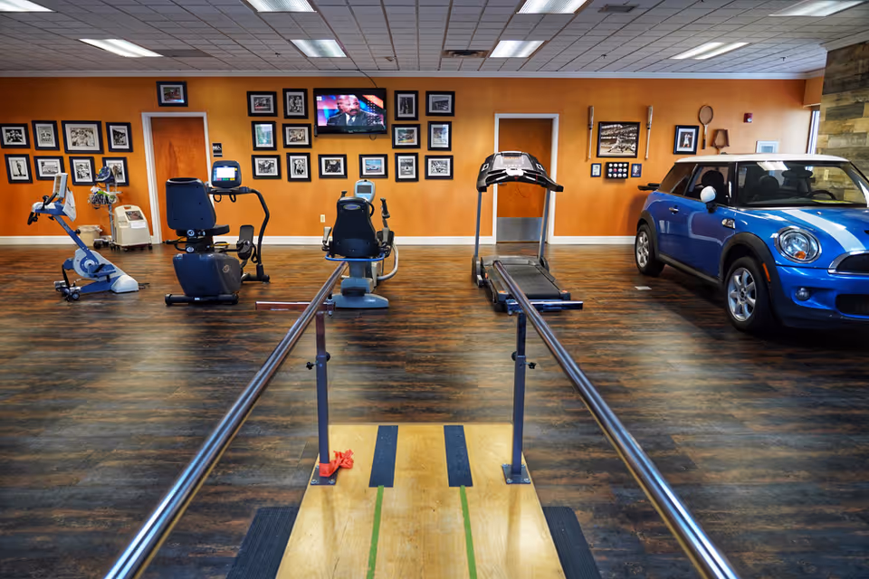 Interior view of a rehabilitation center gym with exercise equipment including stationary bikes, a treadmill, parallel bars for walking support, and a blue Mini Cooper car. The walls are decorated with framed pictures and a television is mounted on the orange wall.