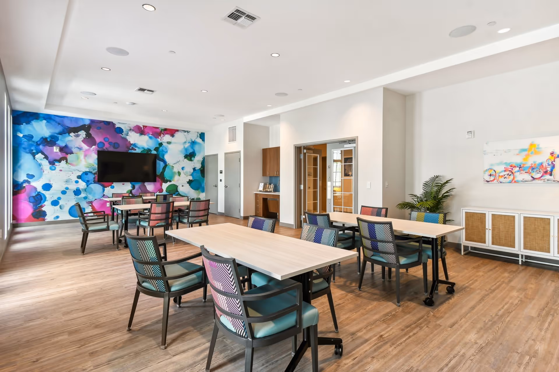 A bright and modern common area in a senior living facility with multiple tables and colorful chairs arranged on a wooden floor. The room features a large abstract mural with blue, purple, and pink hues on one wall, a flat-screen TV mounted on the mural wall, and a sideboard with a colorful painting and a potted plant. There are recessed ceiling lights and a doorway leading to another room.
